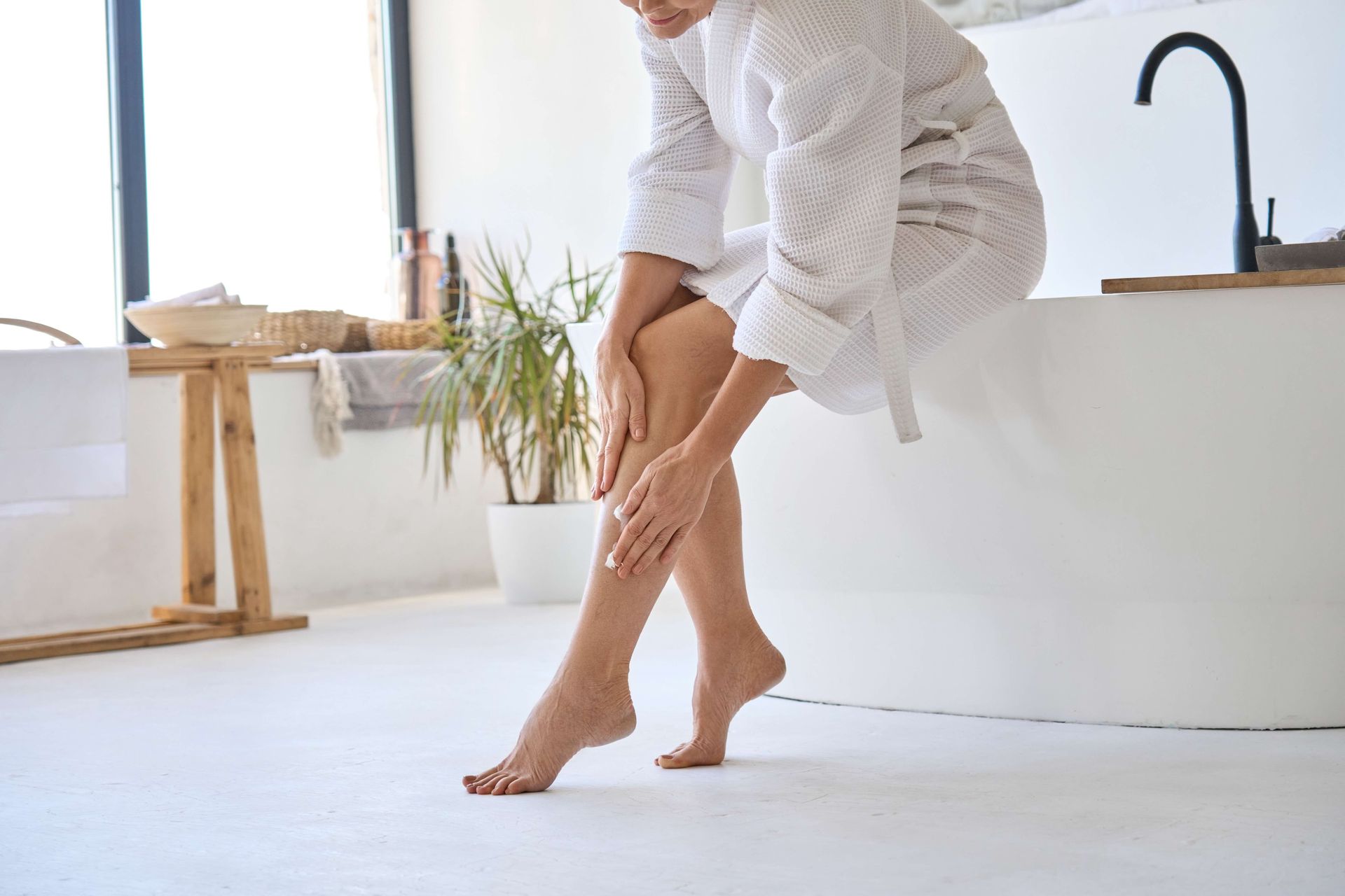 a woman in a bathrobe is sitting on a bathtub and applying lotion to her legs .