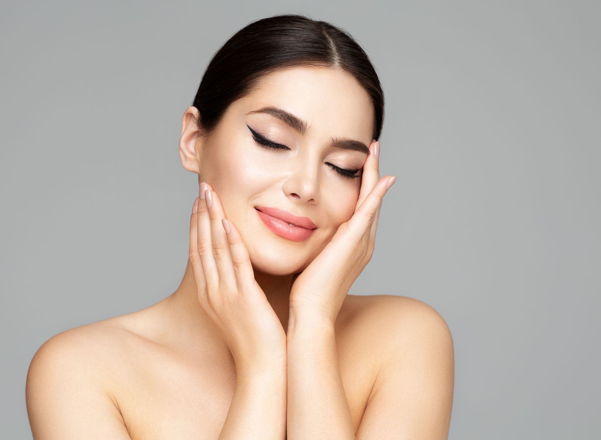 Smiling woman with eyes closed gently touching her cheeks against a gray background