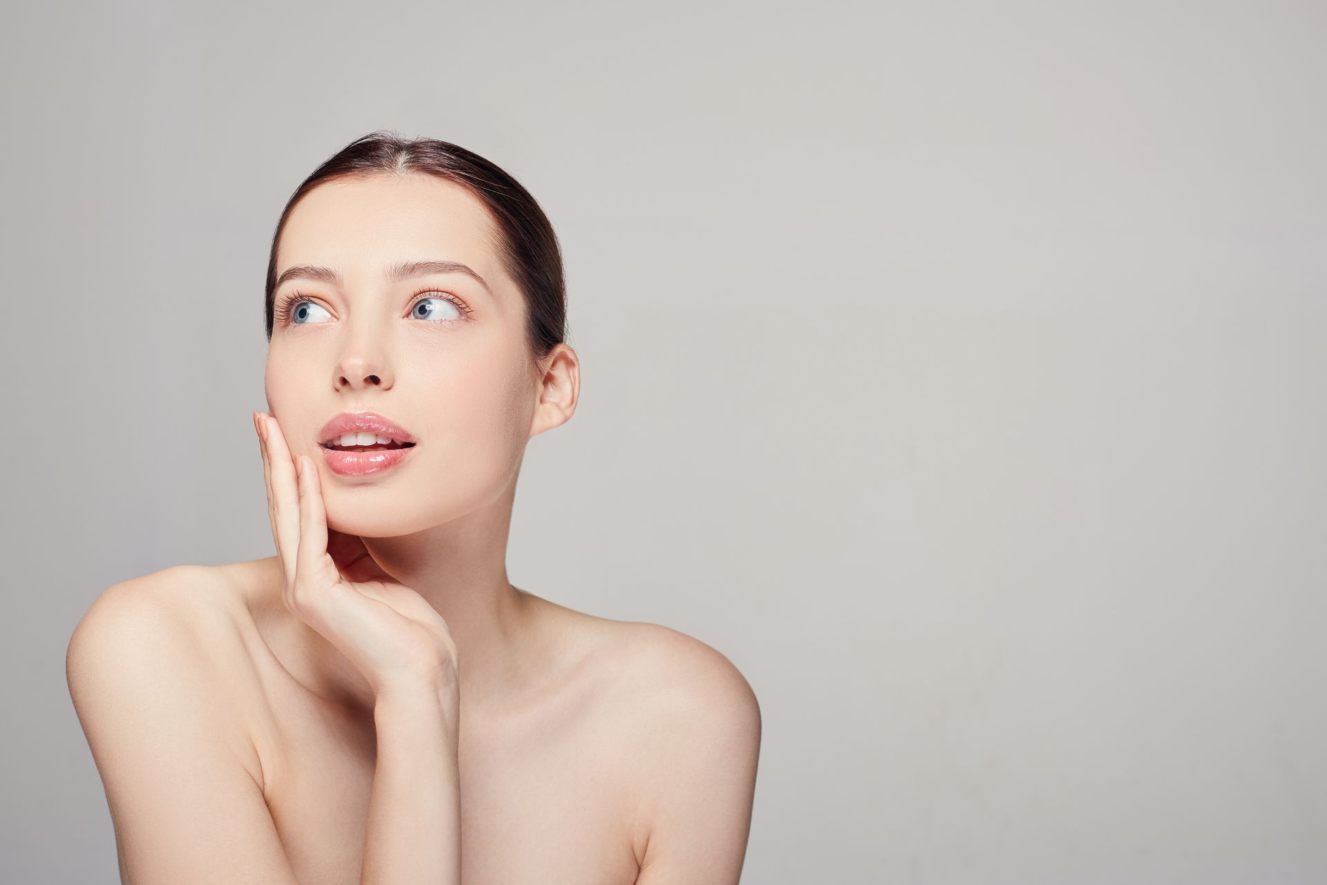Woman with fair skin looking up, touching face, against a gray background.