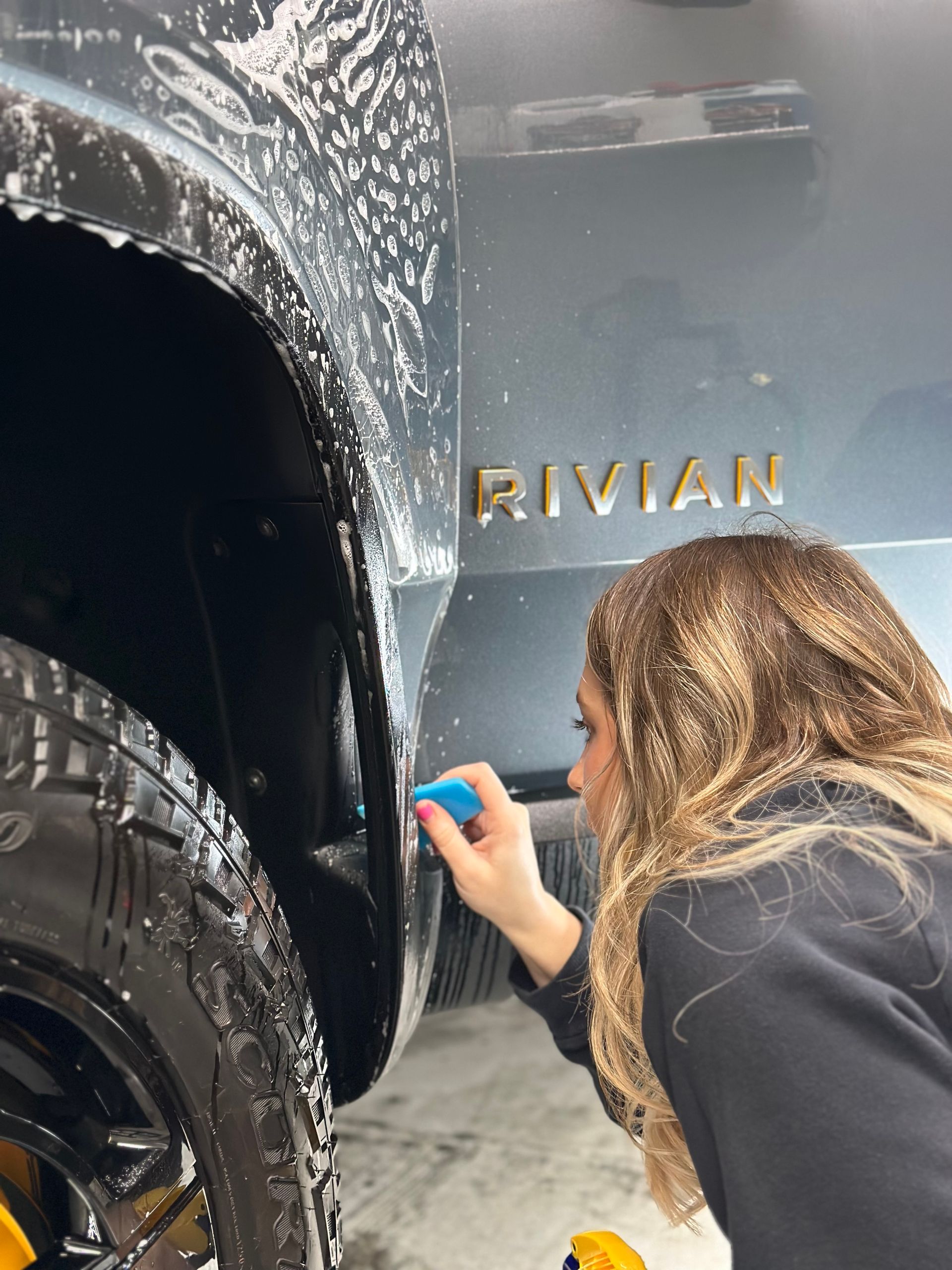 A woman is cleaning the fender of a car with a sponge.