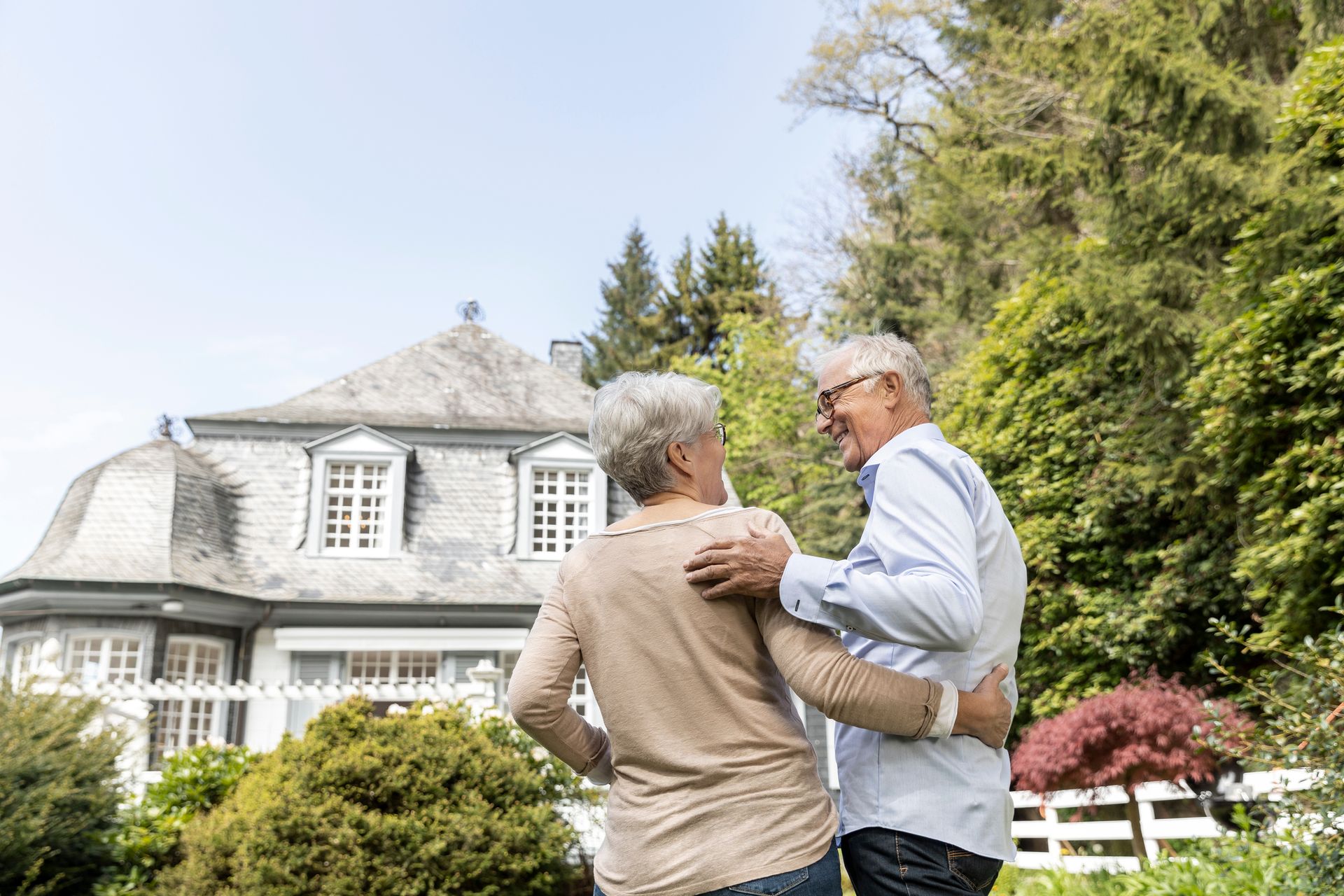 An elderly couple is standing in front of a large house.