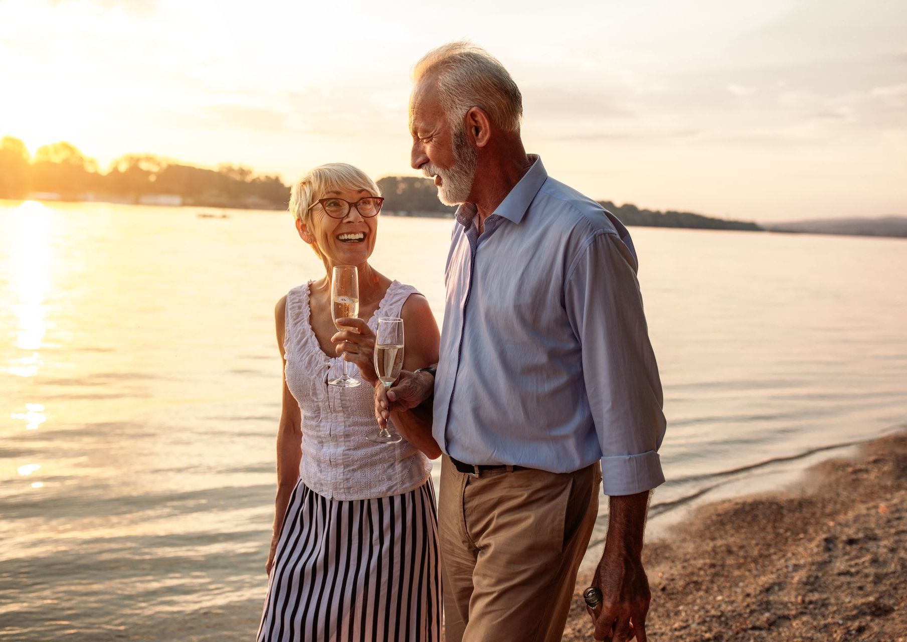 An elderly couple is walking on the beach holding champagne glasses.