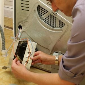 Person fixing the back of a dryer, holding wires. Silver dryer back, white and colored wires.