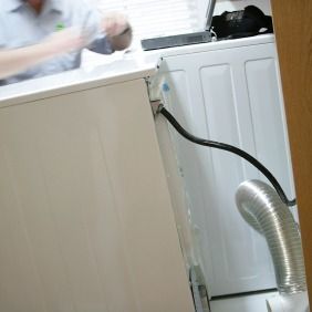 Person near a white dryer with a black power cord and silver exhaust hose.