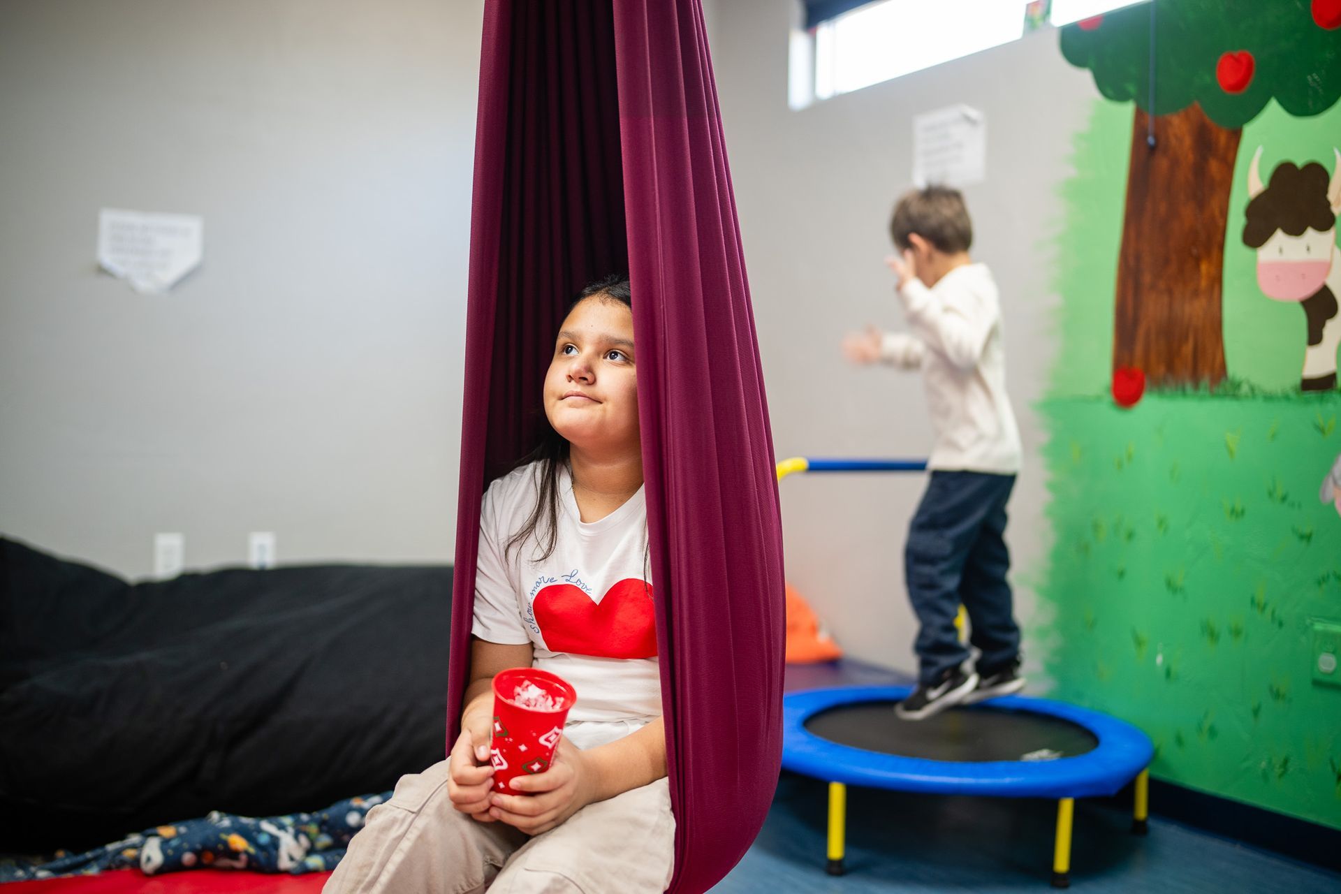 A girl is sitting in a hammock while a boy is jumping on a trampoline.