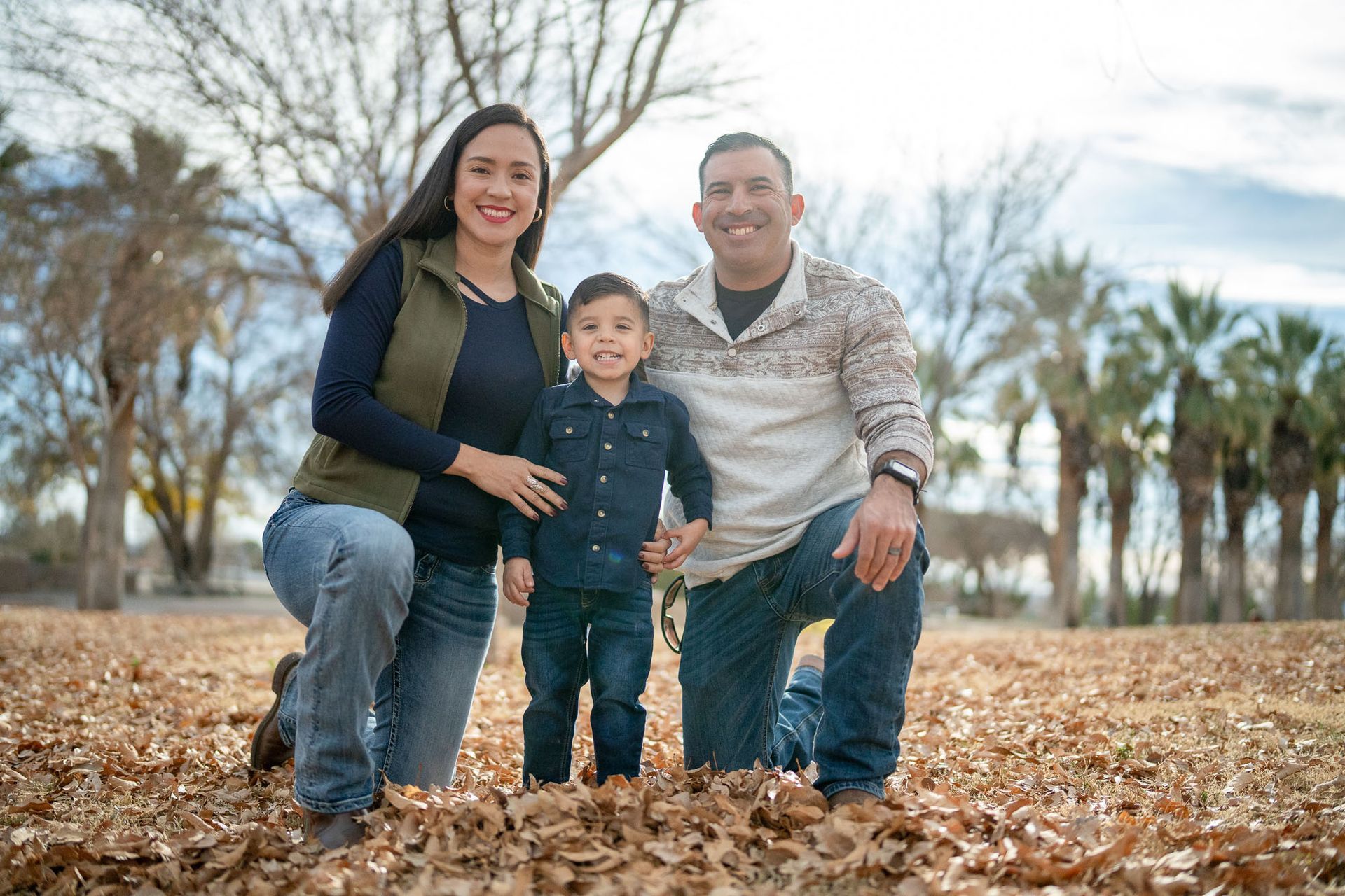 Smiling family of three posing together in a park surrounded by autumn leaves.