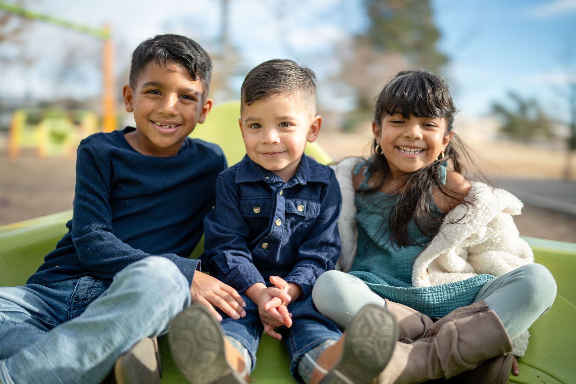 Three smiling children sitting together at a playground representing the inclusive support at Mariposa Autism Service Center.