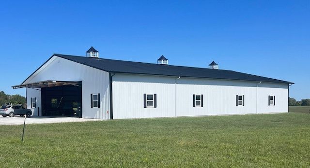A large white barn with a black roof is sitting in the middle of a grassy field.