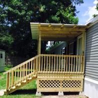 A newly constructed wooden porch with stairs and lattice skirting attached to the side of a grey siding mobile home.