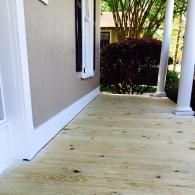 A newly installed light-wood porch floor extends along the side of a house with white columns and trim.