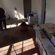 A worker stands in a room undergoing floor renovations, featuring wood planks, a vacuum, and construction supplies.