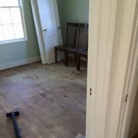 An unfinished room with light-colored flooring, two wooden chairs, a leaning door, and a vacuum handle.