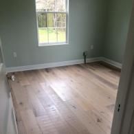 An empty room with pale green walls, light wood flooring, a white window frame, and white baseboards.