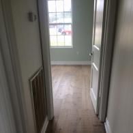 A view down a hallway with light wood floors leading to an open doorway, revealing a room with a window.