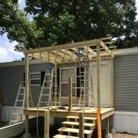 A wooden deck and pergola frame are being built against a mobile home with gray siding, featuring two metal ladders.