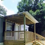 A wooden deck with a covered roof, railings, and a ramp attached to a mobile home under trees.