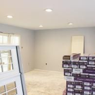 A room under renovation with light gray walls, recessed ceiling lights, a white window frame, and stacked flooring boxes.