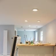 A light-filled room under renovation, featuring recessed ceiling lights, pale blue walls, and a wooden countertop.