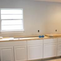 A bright, unfinished kitchen featuring white cabinets and a light wood countertop under a single window.