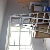 A silver step ladder stands in a room undergoing renovation, positioned in front of a window with paint supplies nearby.