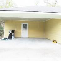 A covered concrete carport attached to a yellow house with a central white door and a large piece of equipment on the left.