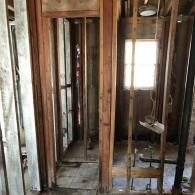 Construction site of an interior room with exposed wooden wall studs, plumbing pipes, and a window in the background.