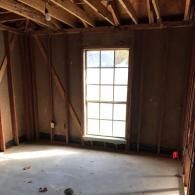 An interior view of a room under construction featuring wooden wall studs, an unfinished floor, and a grid-pane window.
