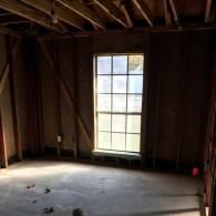 Unfinished room under renovation with wooden wall framing, exposed ceiling joists, and a window letting in natural light.