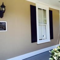 A tan exterior wall with a white-trimmed window, black shutters, a wall-mounted lantern, and a small sign.