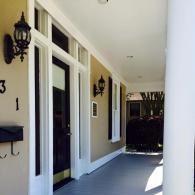 A covered porch with a black front door, a white column, matching wall lamps, and a black mailbox on a tan wall.