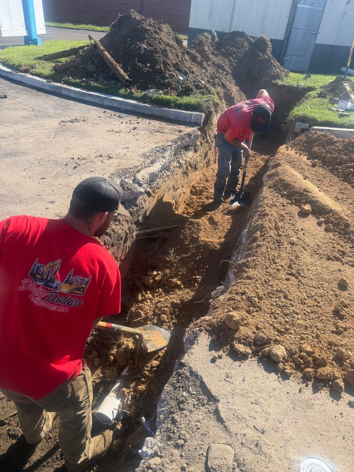 Two workers in red shirts dig a trench near a curb. One is in the foreground, facing away.