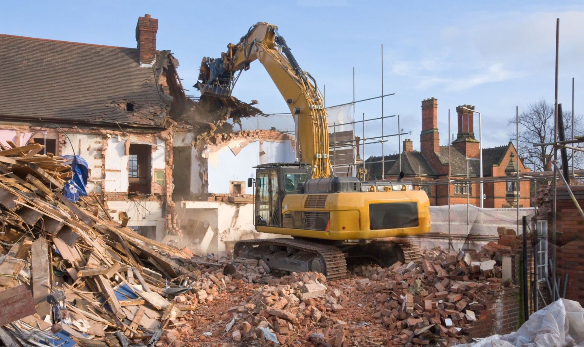 Yellow excavator demolishing a building; debris pile in foreground, other houses in the background.