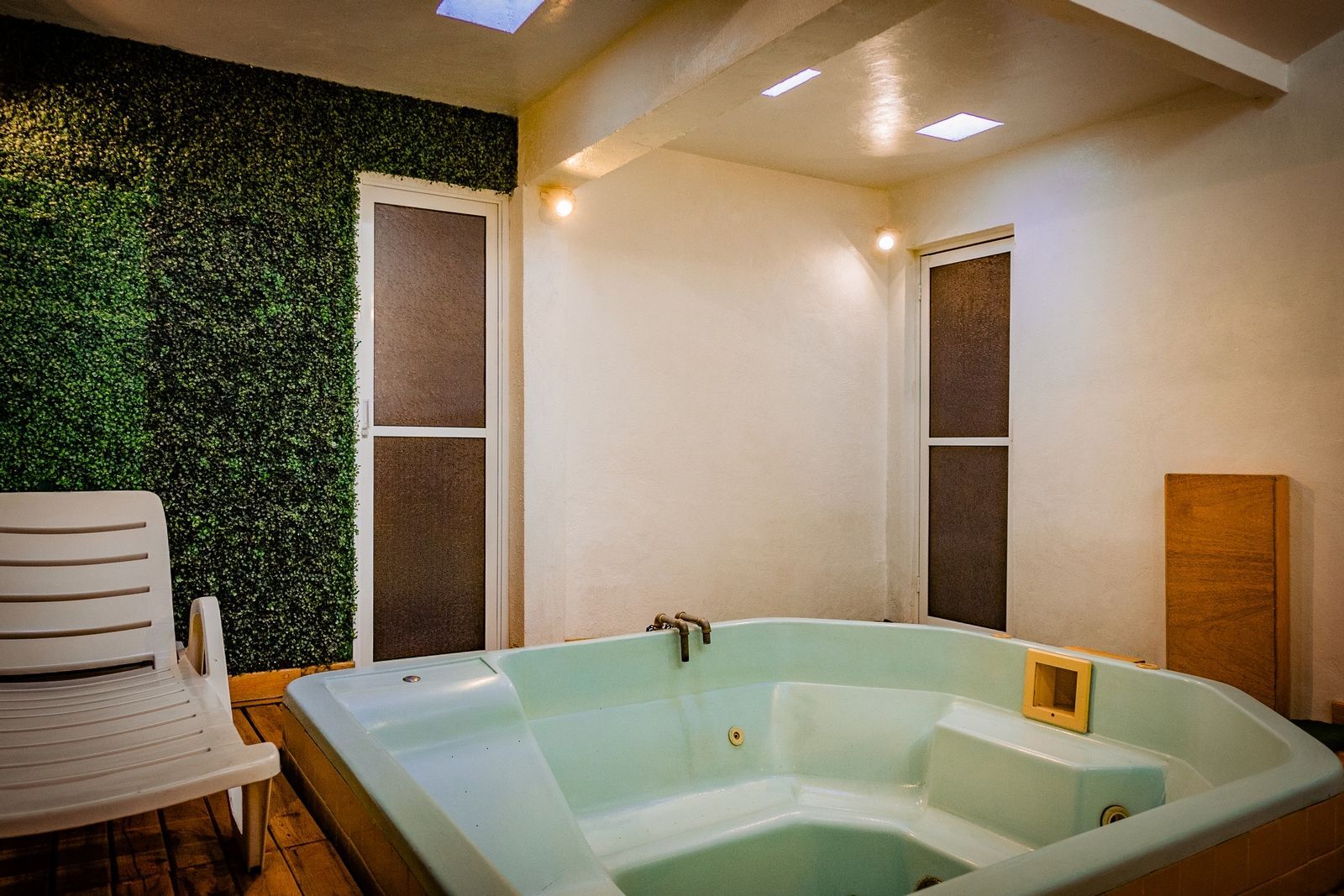 Un jacuzzi en una habitación con una pared verde artificial, paredes blancas y un sillón.