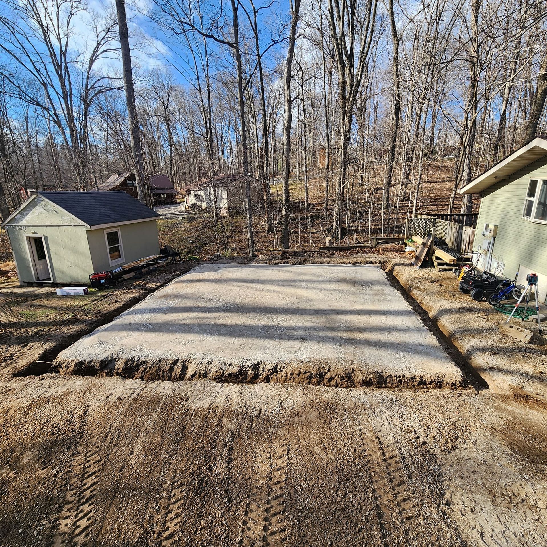 A rectangular gravel pad, surrounded by dirt, is prepped for construction near two small buildings in a wooded area.