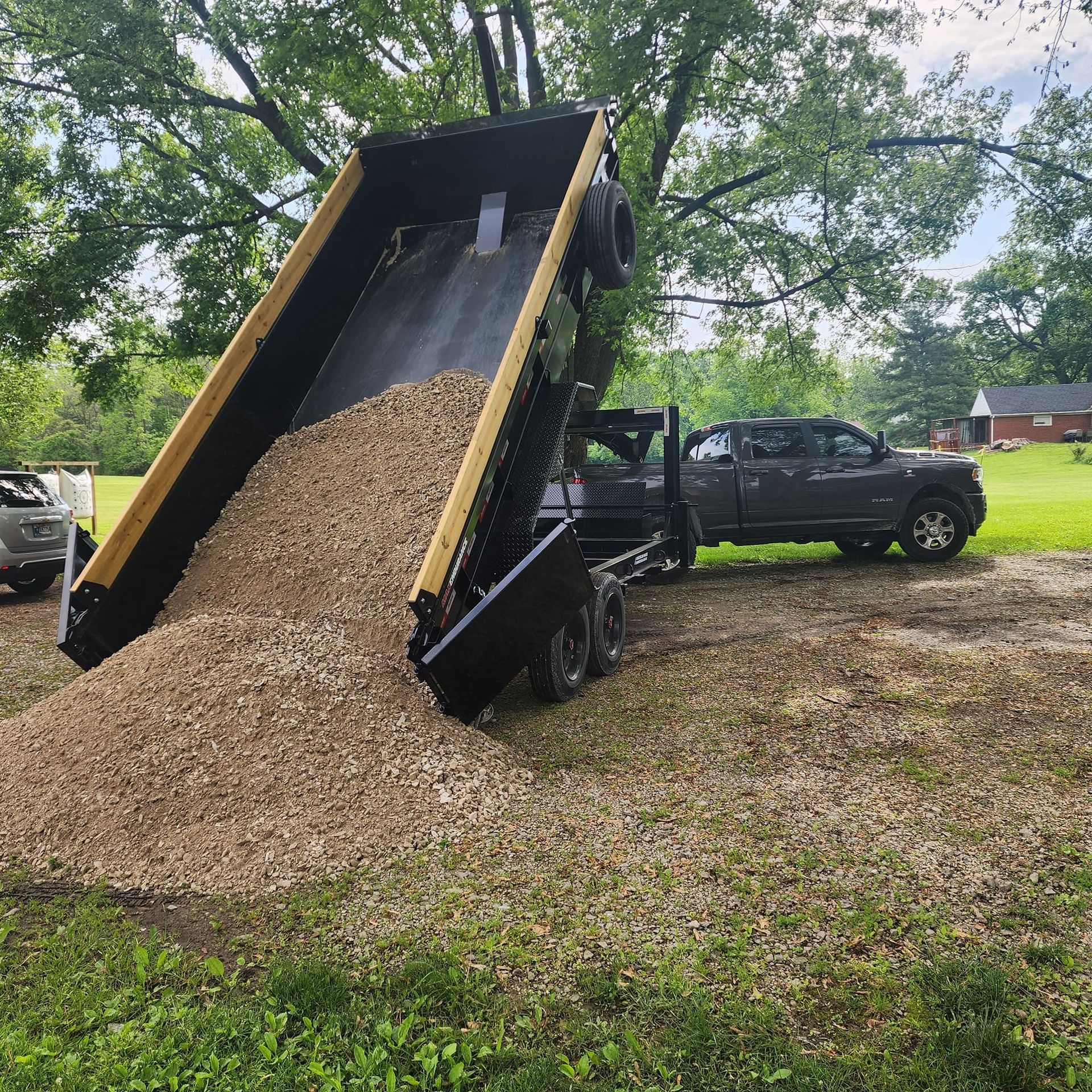 Dump truck trailer dumping gravel on a grassy area, with a pickup truck attached.