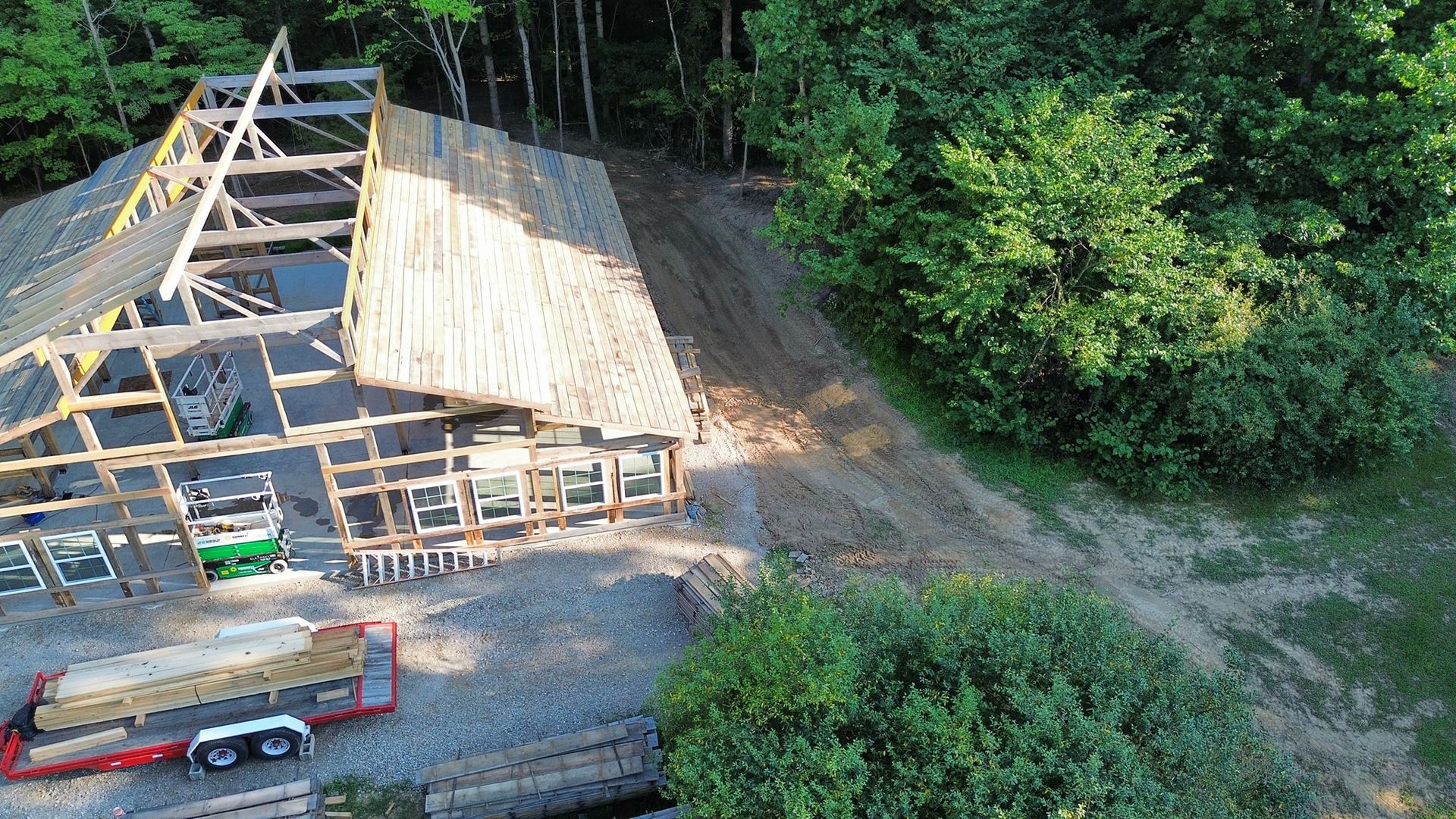 Construction site: partially built wood-framed house in a clearing, surrounded by trees and a dirt road.