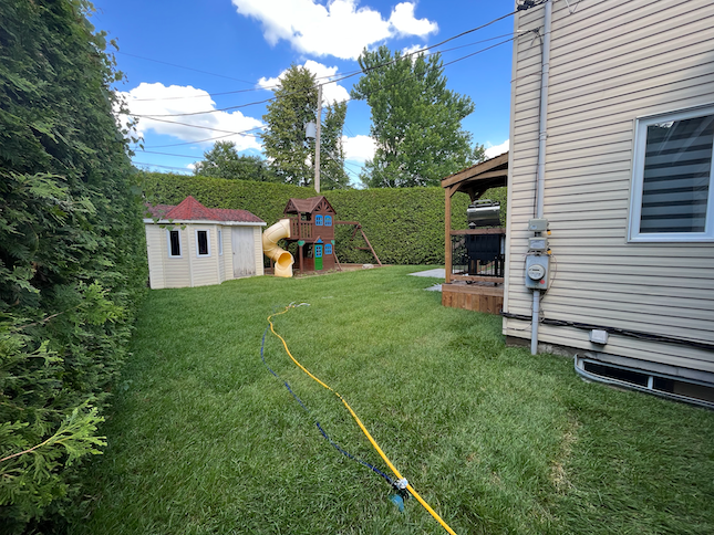 Un jardin avec une petite cabane de jeux, une balançoire pour enfants avec un toboggan jaune et une terrasse en bois avec un barbecue.