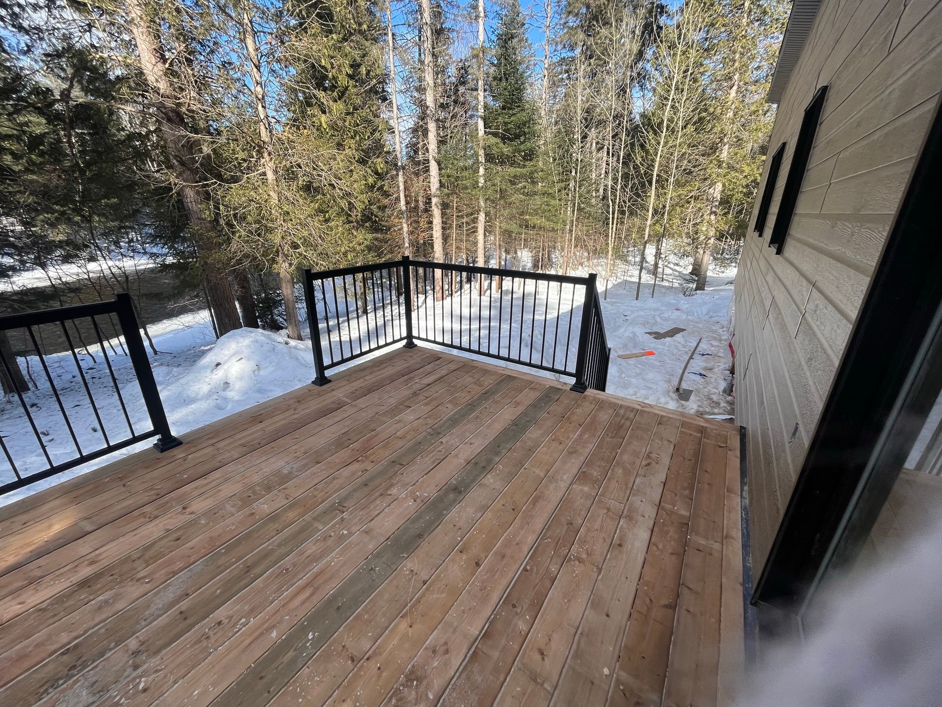 Une terrasse en bois avec une balustrade en métal est entourée de neige et d'arbres.