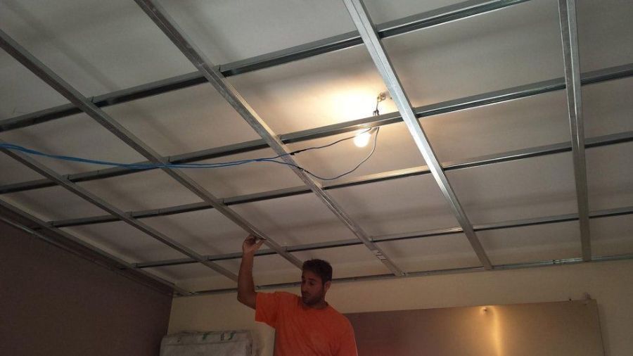 Uomo che installa un controsoffitto con griglia metallica e isolamento. Le luci pendono, la stanza ha pareti marroni e bianche.