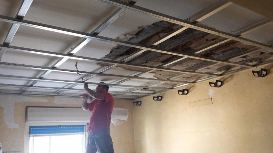 Uomo che installa un controsoffitto. Il soffitto è parzialmente rimosso, rivelando travi in ​​legno. Pareti gialle.