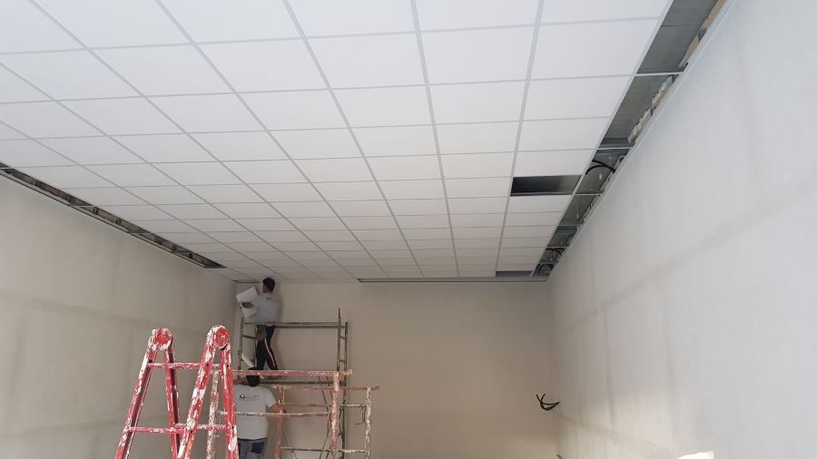 Operai che installano un controsoffitto in una stanza dalle pareti bianche, uno su un'impalcatura, l'altro su una scala rossa.