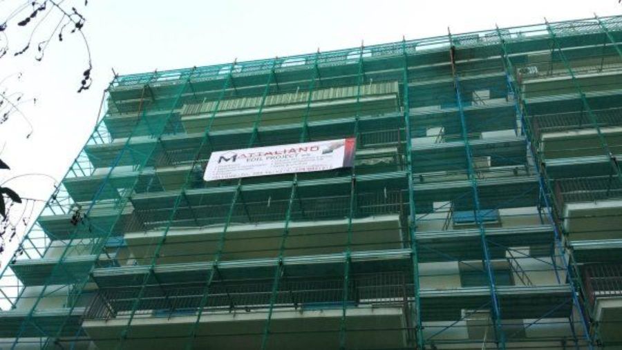 Edificio in fase di ristrutturazione, coperto da reti di sicurezza verdi e impalcature; striscione con il logo aziendale.