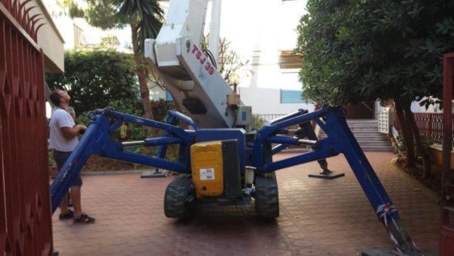 Piattaforma aerea ragno blu e gialla con braccio sollevato, in un cortile. Sono presenti due persone.