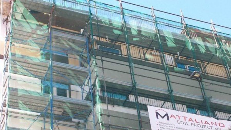 Esterno dell'edificio in costruzione, coperto da impalcature e reti di sicurezza. È visibile lo striscione 