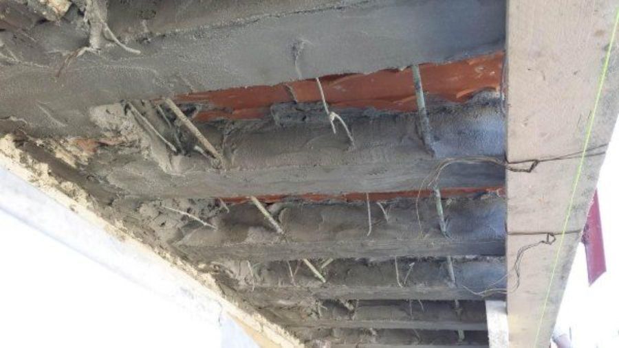 Soffitto in cemento danneggiato che espone tondini di ferro e mattoni. Cemento grigio, con dettagli marroni e verdi.