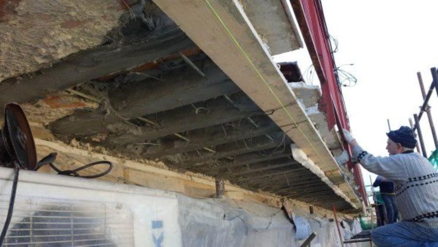 Un operaio edile misura travi di legno sotto una sporgenza di cemento, con le impalcature visibili.