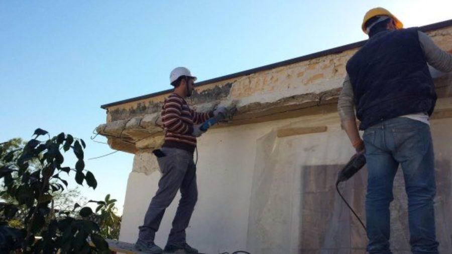 Due operai edili su un tetto lavorano con utensili elettrici sotto un cielo azzurro e terso.