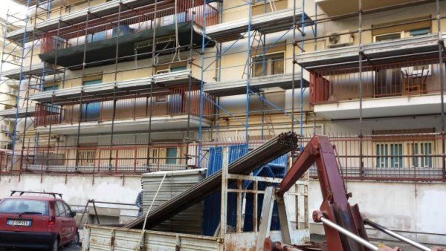 Edificio con impalcature; lavori di costruzione. Auto e camion rossi davanti.