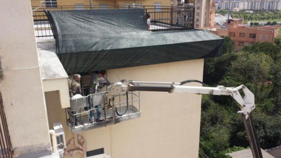 Operai su una piattaforma aerea al lavoro su un edificio; sul tetto c'è un telo nero e le pareti dell'edificio sono beige.