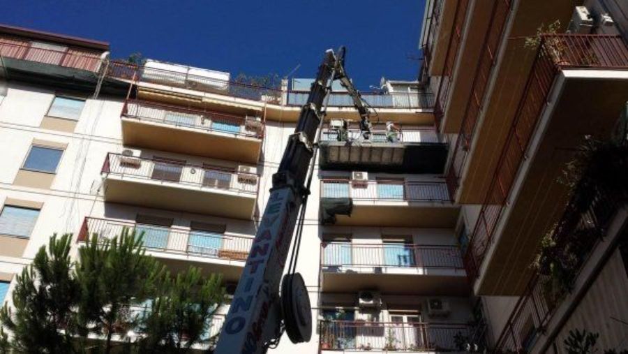 Una gru solleva una piattaforma verso un condominio a più piani, in una giornata di sole.